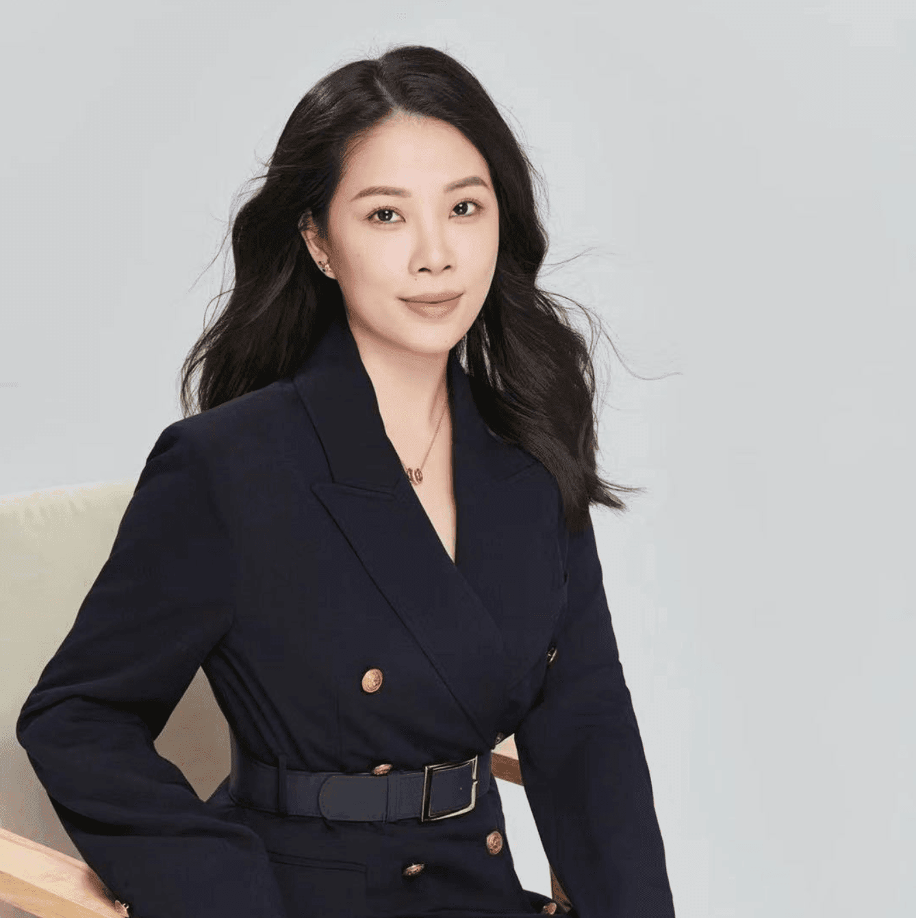 Woman with long black hair in a dark blazer sitting on a chair against a light gray background.